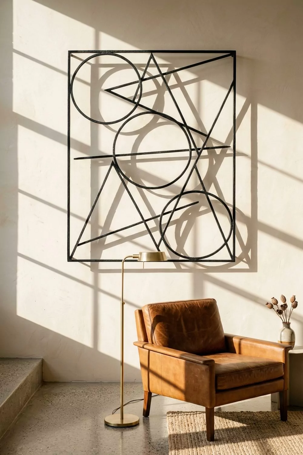 Large open black iron geometric wall sculpture with circular and angular shapes mounted on a warm white plaster wall above a leather mid-century lounge chair in a modern living room