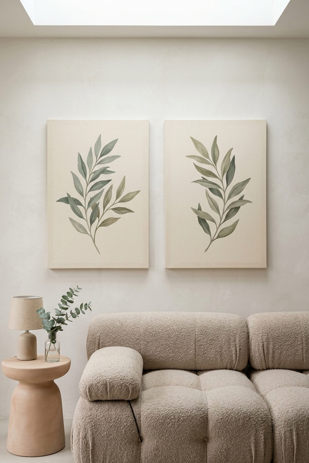 Pair of vertical botanical canvas prints featuring muted sage and olive leaf paintings hung symmetrically above a beige bouclé sofa in an airy neutral living room