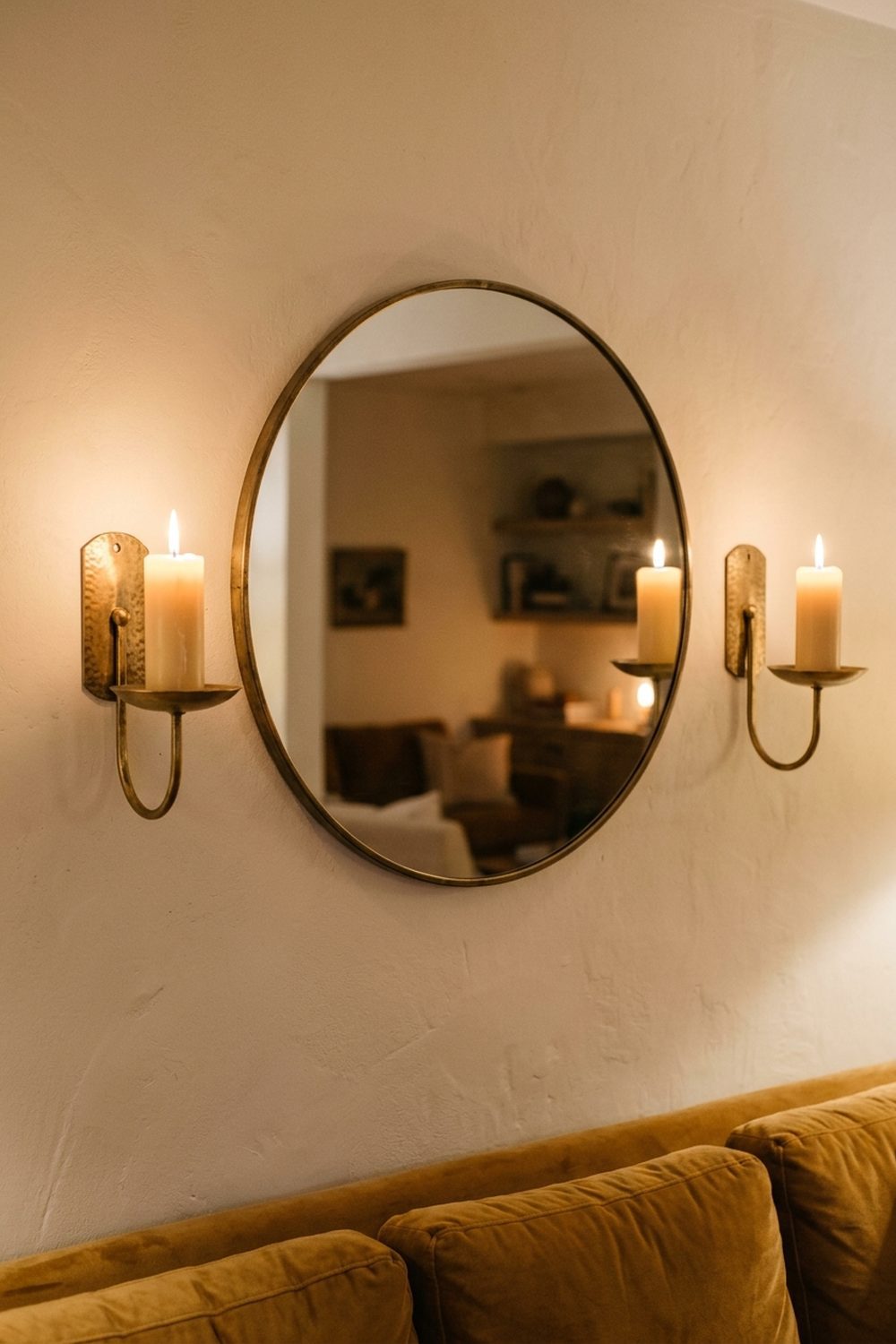 Pair of antique brushed brass wall candle sconces with pillar candles flanking a large round mirror on a soft cream wall in an elegant living room