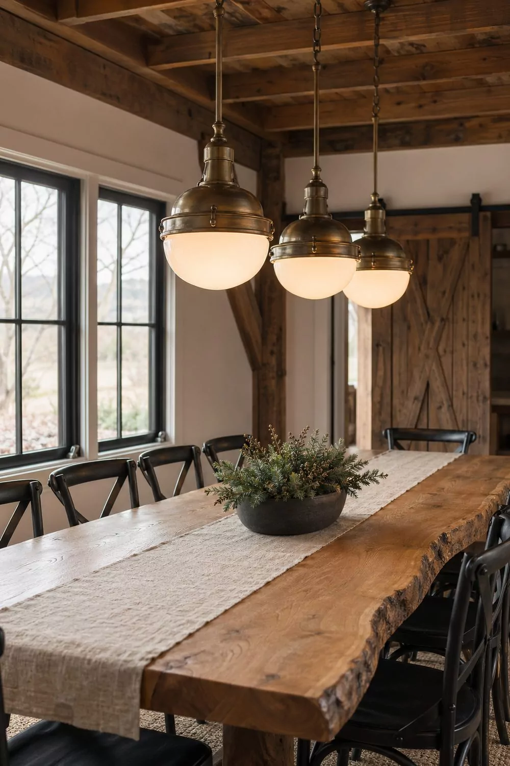 Industrial pendant barn dining area with three aged brass opal glass pendants over a live edge oak table, black metal cross back chairs, linen runner, ceramic bowl with greens