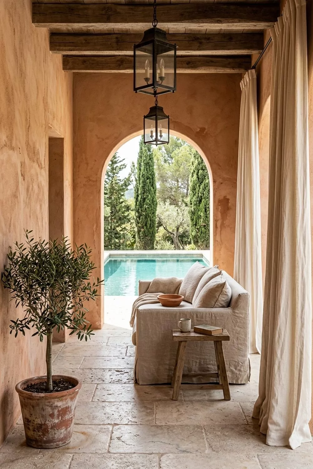 Mediterranean stucco pool cabana with terracotta plaster walls, arched opening framing a pool and cypress trees, iron lanterns and linen sofa