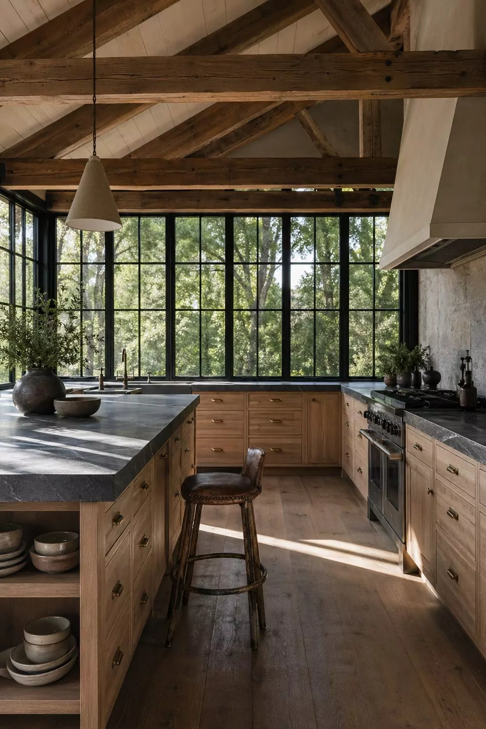 Modern barn kitchen with tall black-framed steel windows, soapstone island, white oak cabinetry with brushed brass pulls, exposed wood ceiling beams, vintage leather stool