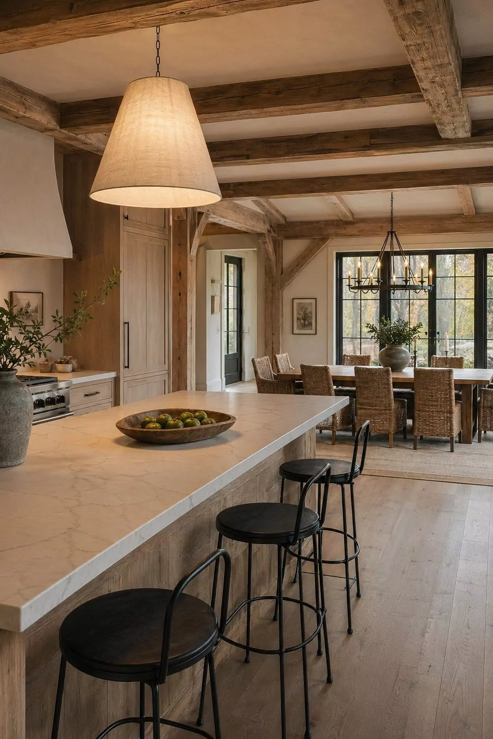 Open concept barn dining kitchen with honed quartz island, linen pendant, white oak cabinetry, exposed wood ceiling beams, oak dining table with rattan chairs, wrought iron chandelier, black framed windows