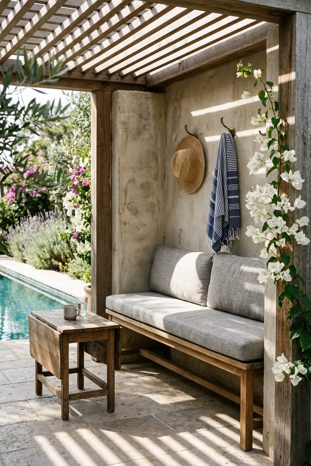 Small pergola pool cabana nook with slim bench, linen cushions, brass hooks with straw hat and striped towel, bougainvillea and pool beyond