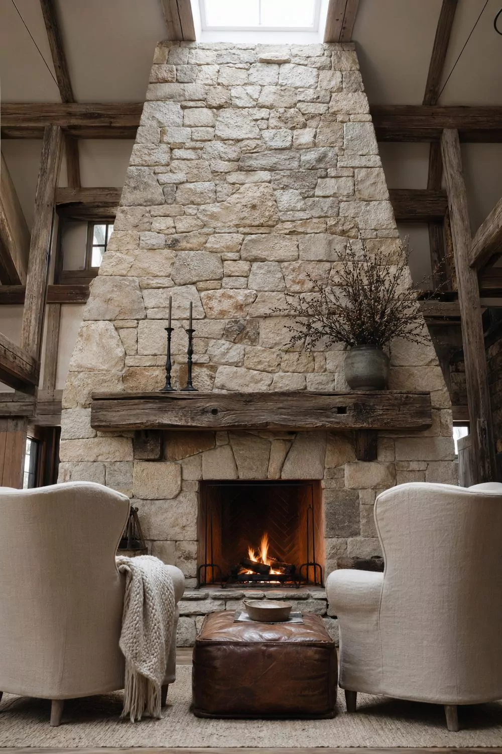 Floor to ceiling fieldstone fireplace barn great room with reclaimed wood mantel, cream linen wingback chairs, leather ottoman, cast iron candlesticks