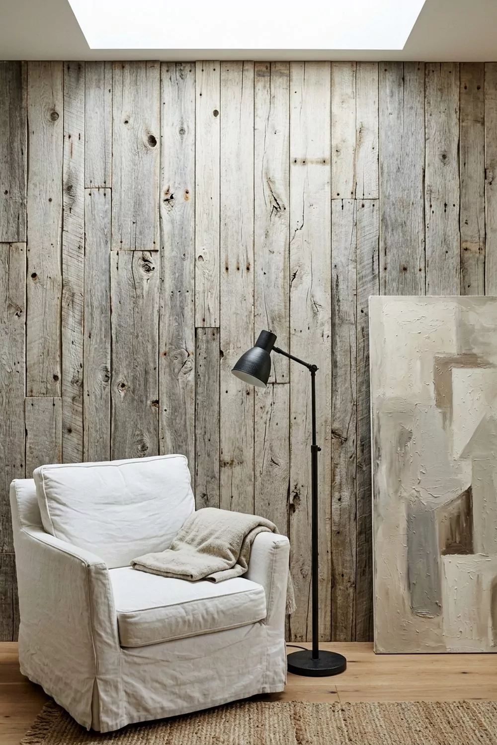 Weathered reclaimed driftwood plank coastal accent wall with sun bleached grey planks of varying widths, natural knots and cracks, behind a slipcovered armchair and black iron floor lamp