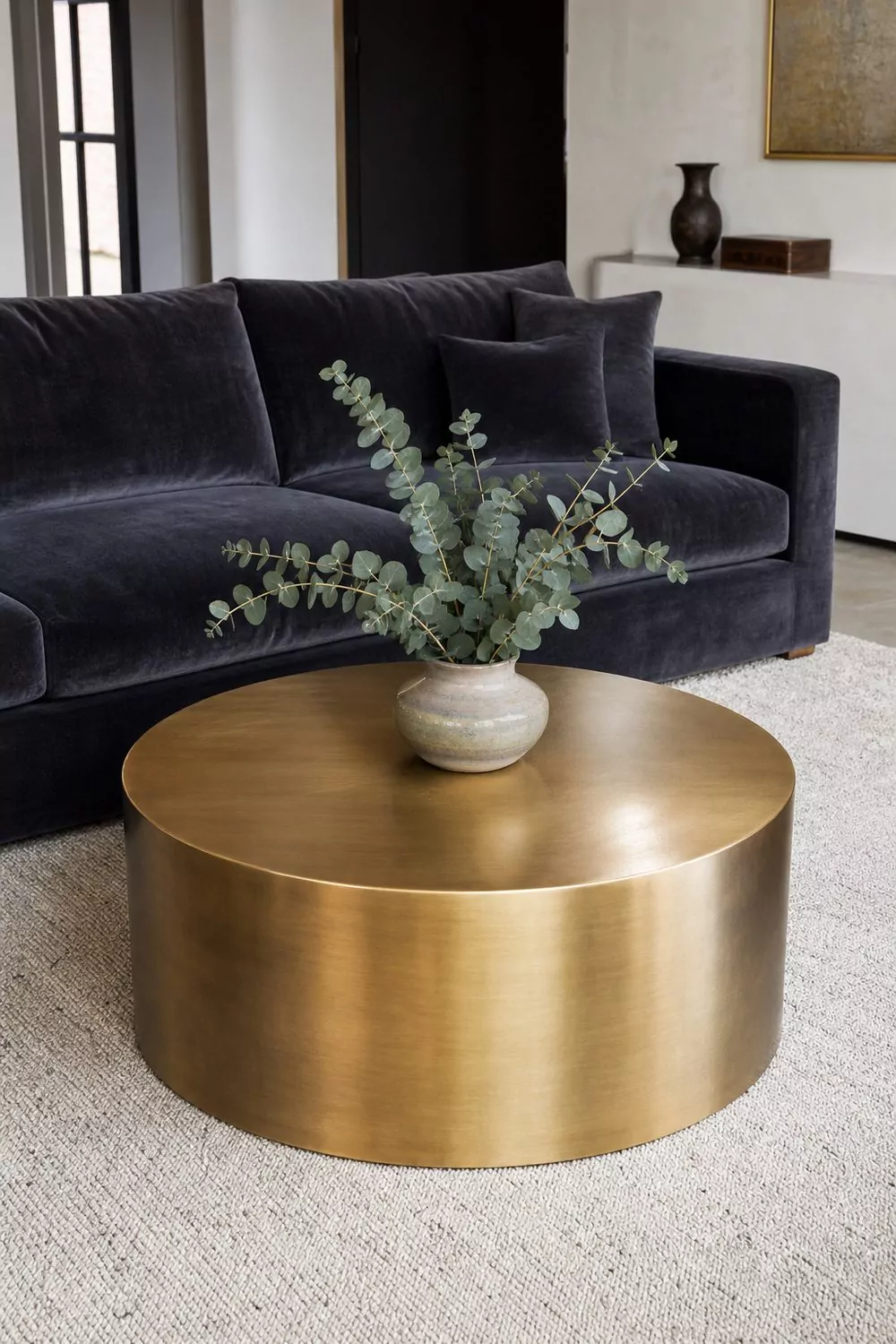 Round brushed antique brass coffee table with eucalyptus stems in a ceramic vase placed in front of a charcoal velvet sofa on an ivory wool rug
