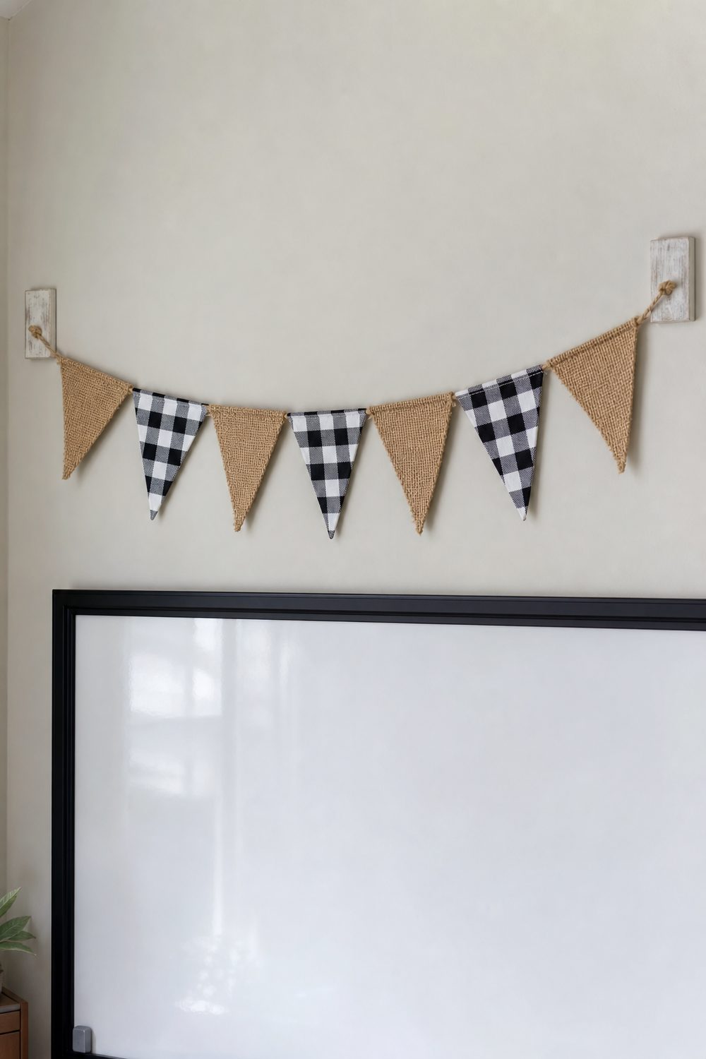 Farmhouse classroom welcome banner made of alternating burlap and black and white buffalo check fabric pennants strung on natural twine across a soft white wall above a whiteboard, bright midday diffused light