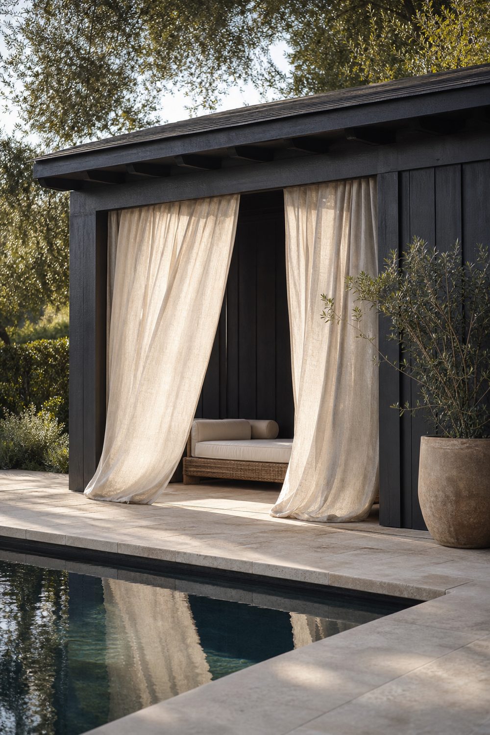 Small charcoal painted pool cabana with cream linen curtains billowing at the open front, rattan lounger inside, light limestone pool deck, soft morning light