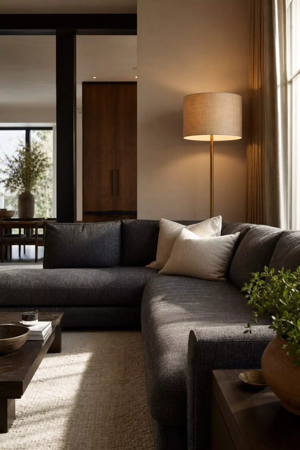 Deep charcoal grey L-shaped sectional sofa anchored by a slim brushed brass floor lamp with warm linen drum shade in a modern living room
