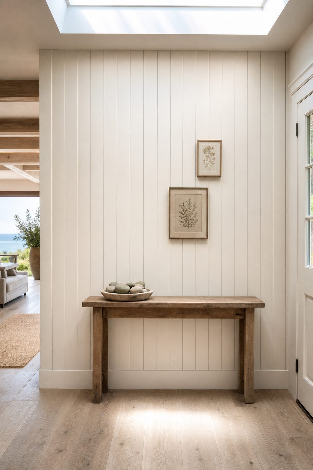 Vertical warm white shiplap accent wall with two small framed botanical prints, weathered teak bench, and a wood bowl of sea worn stones in a coastal entryway