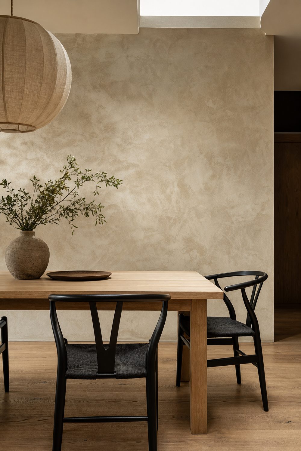 Warm cream lime plaster accent wall with cloud like variation behind a pale oak dining table and matte black wishbone chairs in a Japandi dining room