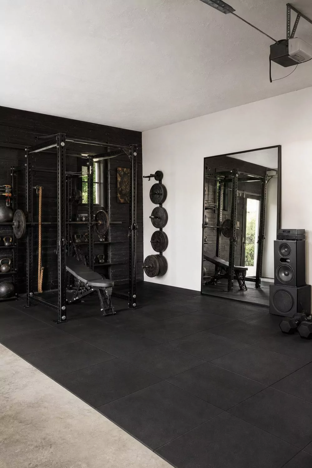 Garage gym man cave with black rubber flooring, wall-mounted weight rack, vertical plate storage and full-length mirror in bright daylight