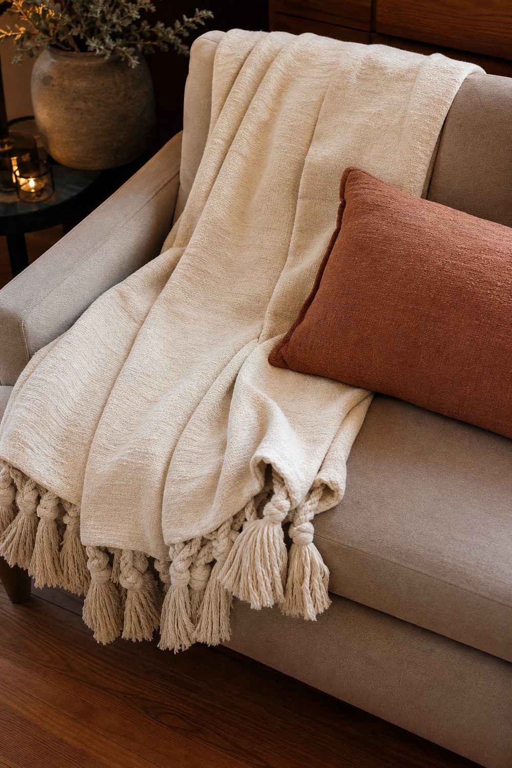 Oversized cream throw blanket with chunky tassels folded across a taupe sofa seat with clay lumbar pillow