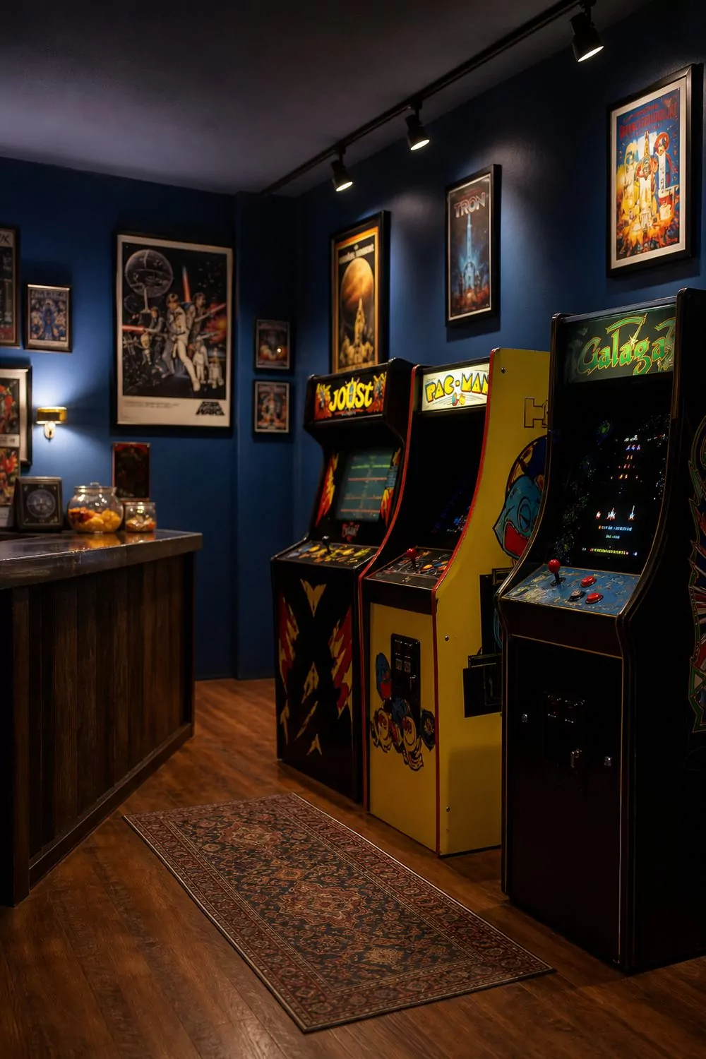 Vintage arcade man cave with three upright cabinets, snack counter, retro posters and deep-blue walls under dim glowing track lighting