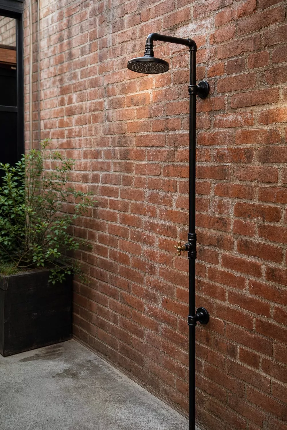 Industrial outdoor shower of black steel pipe with a brass valve on a red brick courtyard wall over concrete, soft morning light