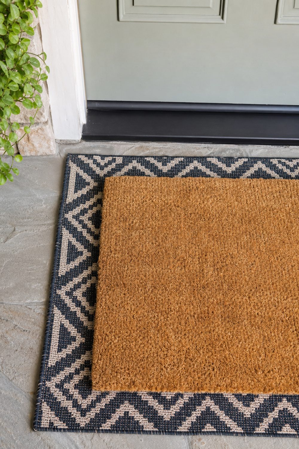 Natural coir doormat layered over a patterned flatweave outdoor rug at a front door threshold, viewed from above in bright midday light