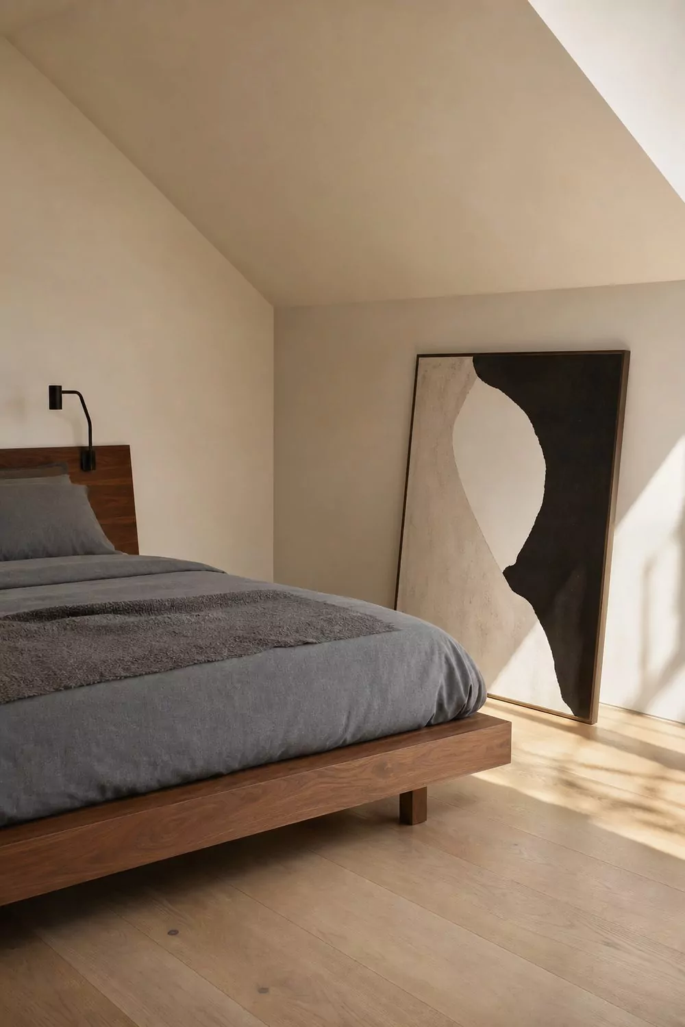 Modern minimalist attic bedroom with a low walnut platform bed, soft white sloped ceiling, monochrome grey bedding and pale oak flooring