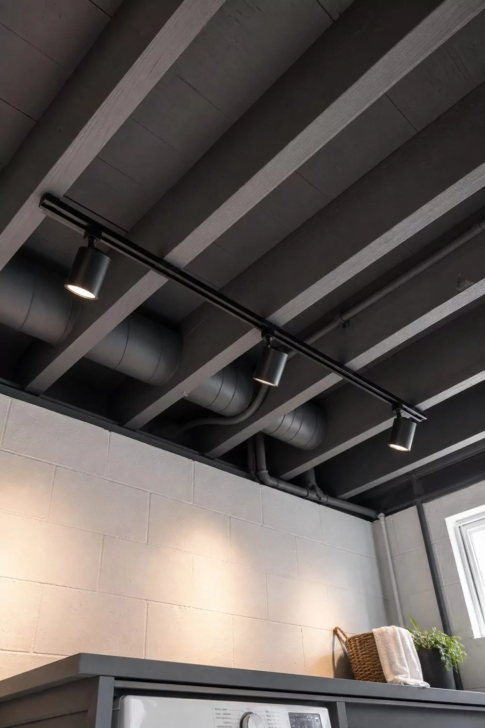 Basement ceiling with exposed joists ductwork and pipes painted uniform charcoal, with a black track light and three adjustable heads