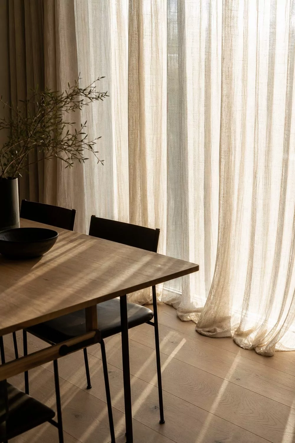 Full height sheer linen curtains filtering daylight behind a pale oak dining table with slim black chairs in a contemporary dining room