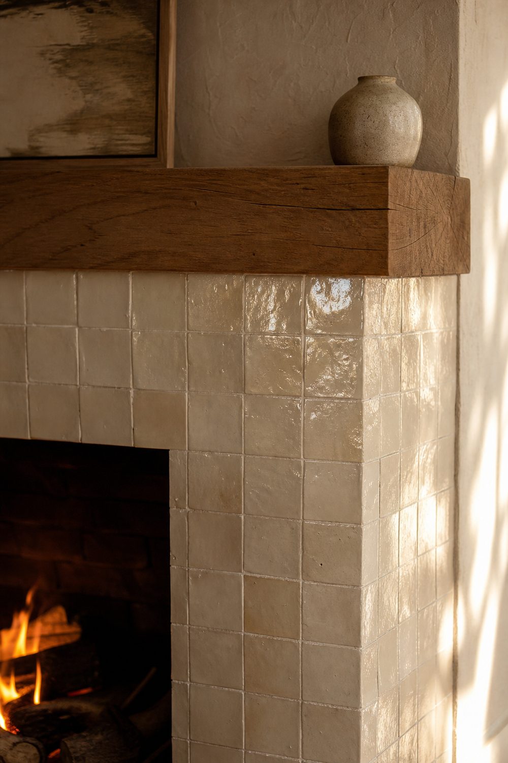 Fireplace surround in handmade cream zellige tile with glossy uneven surface catching firelight beside an oak mantel