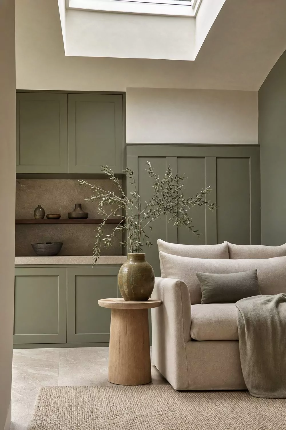 Beige and sage green room with green painted cabinetry, beige linen chair and an olive ceramic vase in bright daylight