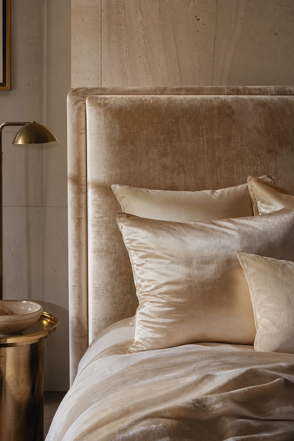 Beige velvet headboard with visible pile sheen styled with champagne silk cushions, brass floor lamp and stone wall behind