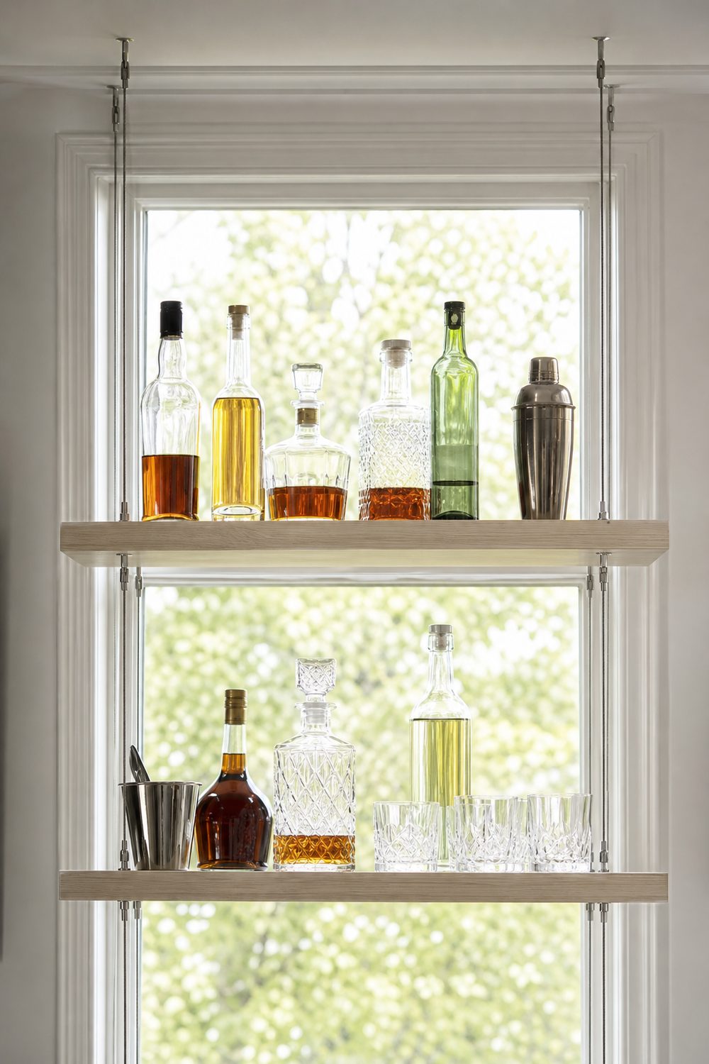 Two slim ash shelves suspended from the ceiling on thin stainless steel cables in front of a bright window with bottles and cut glass tumblers