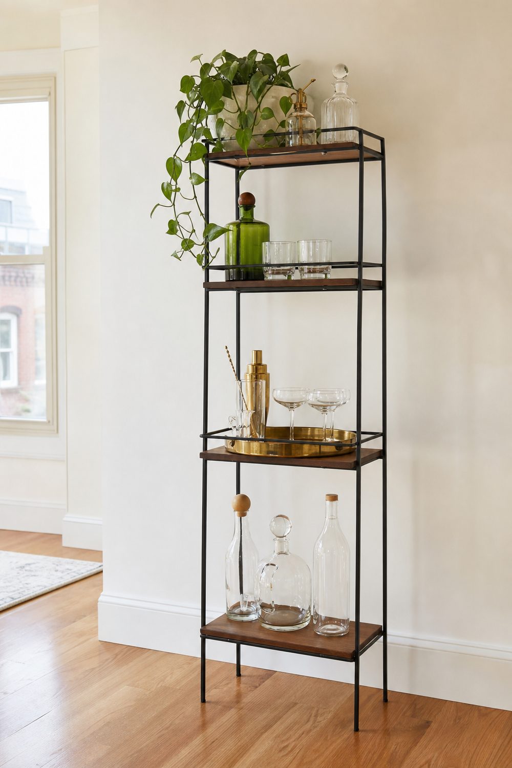 Slim freestanding black metal etagere with four wood tiers used as an apartment bar against a plain white wall with bottles, brass tray and a trailing plant