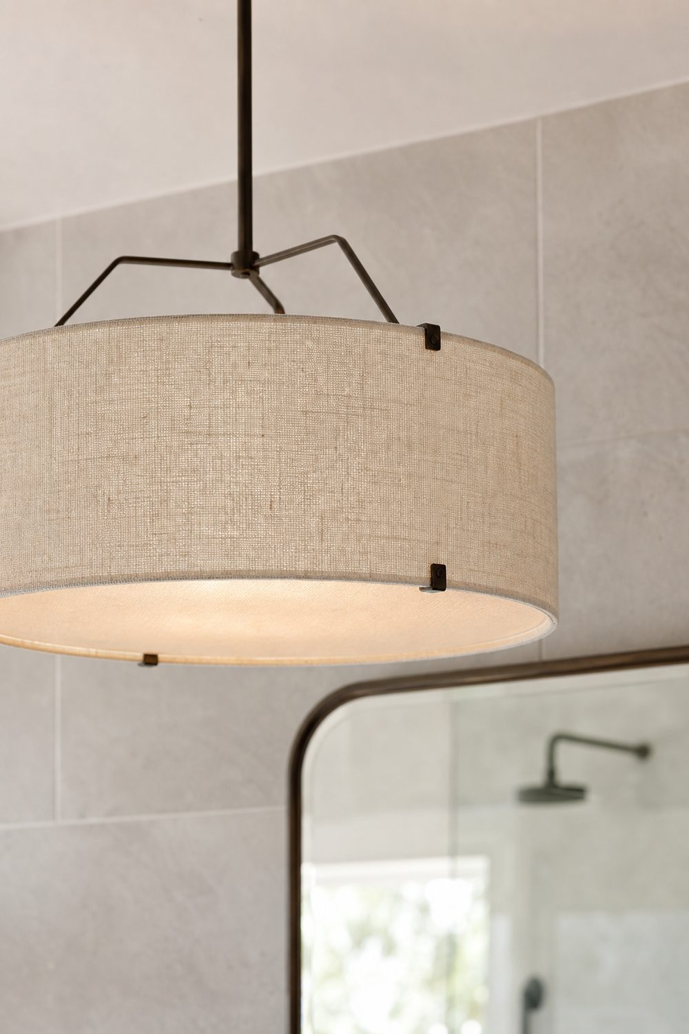Close up of a natural linen drum chandelier with a bronze frame glowing softly against pale grey bathroom wall tile