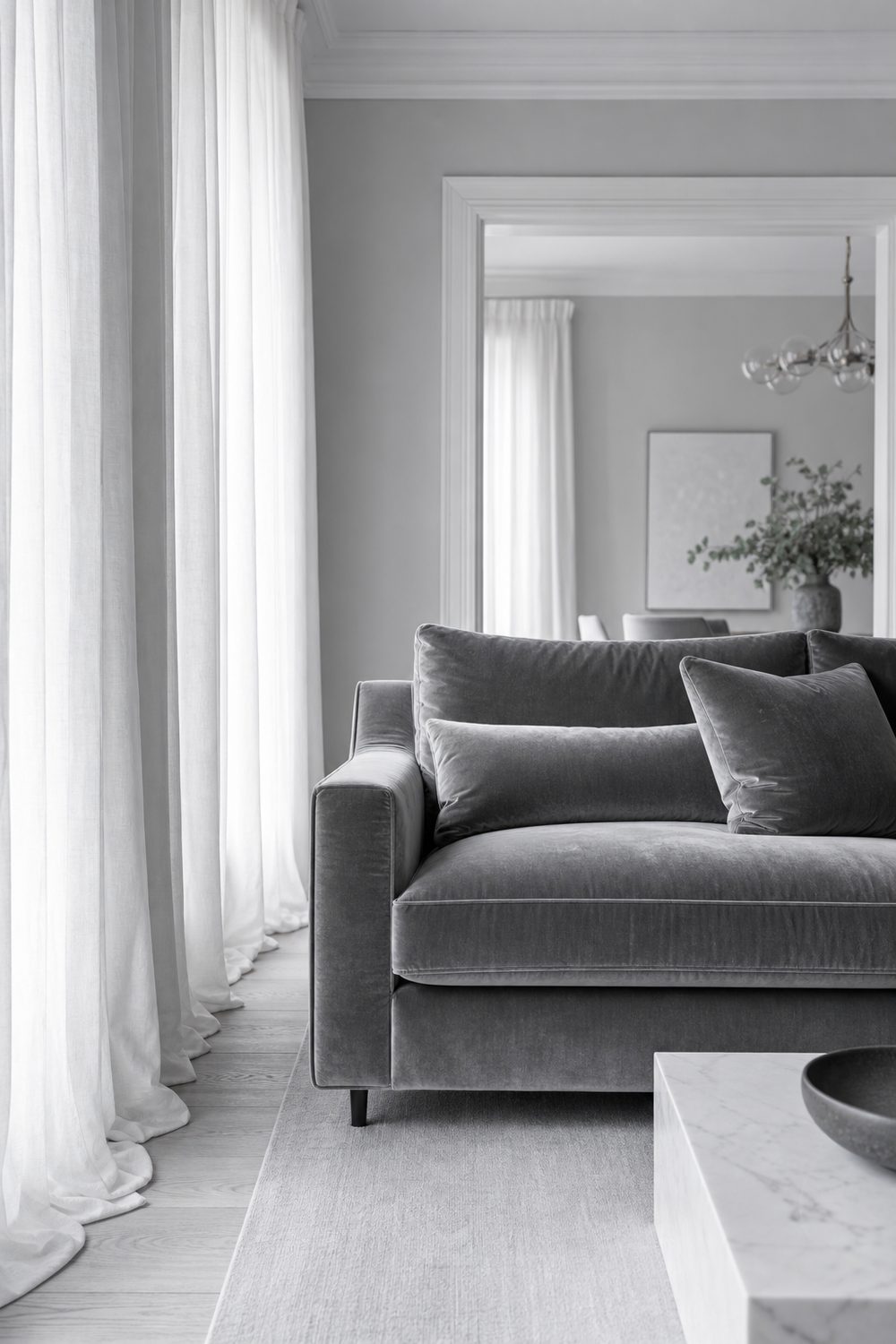 Grey velvet sofa with visible pile beside floor length white linen curtains in a light grey living room