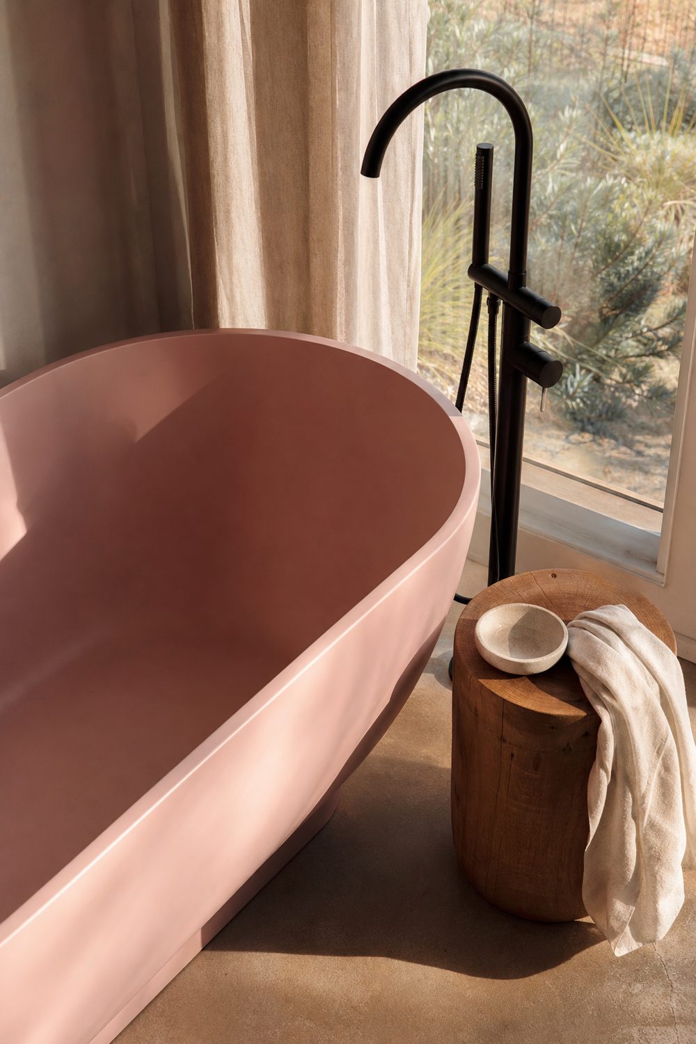 Freestanding soaking tub in millennial pink before a tall window, with a floor mounted matte black tub filler and an oak stool