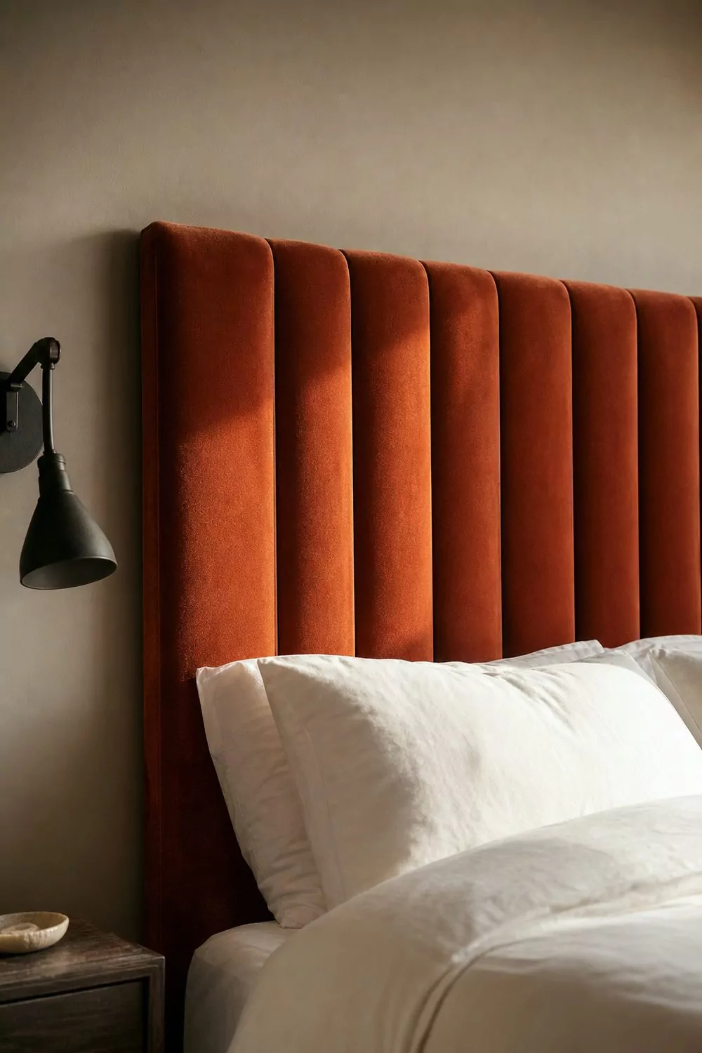 Tall channel tufted rust velvet headboard against a warm greige wall with crisp white bedding and a black metal sconce