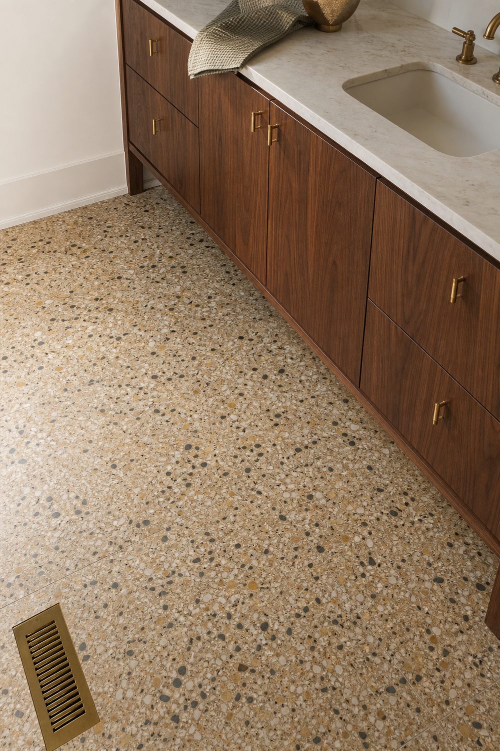 Warm terrazzo floor flecked with ochre and charcoal chips beside a walnut vanity with brass pulls and floor register