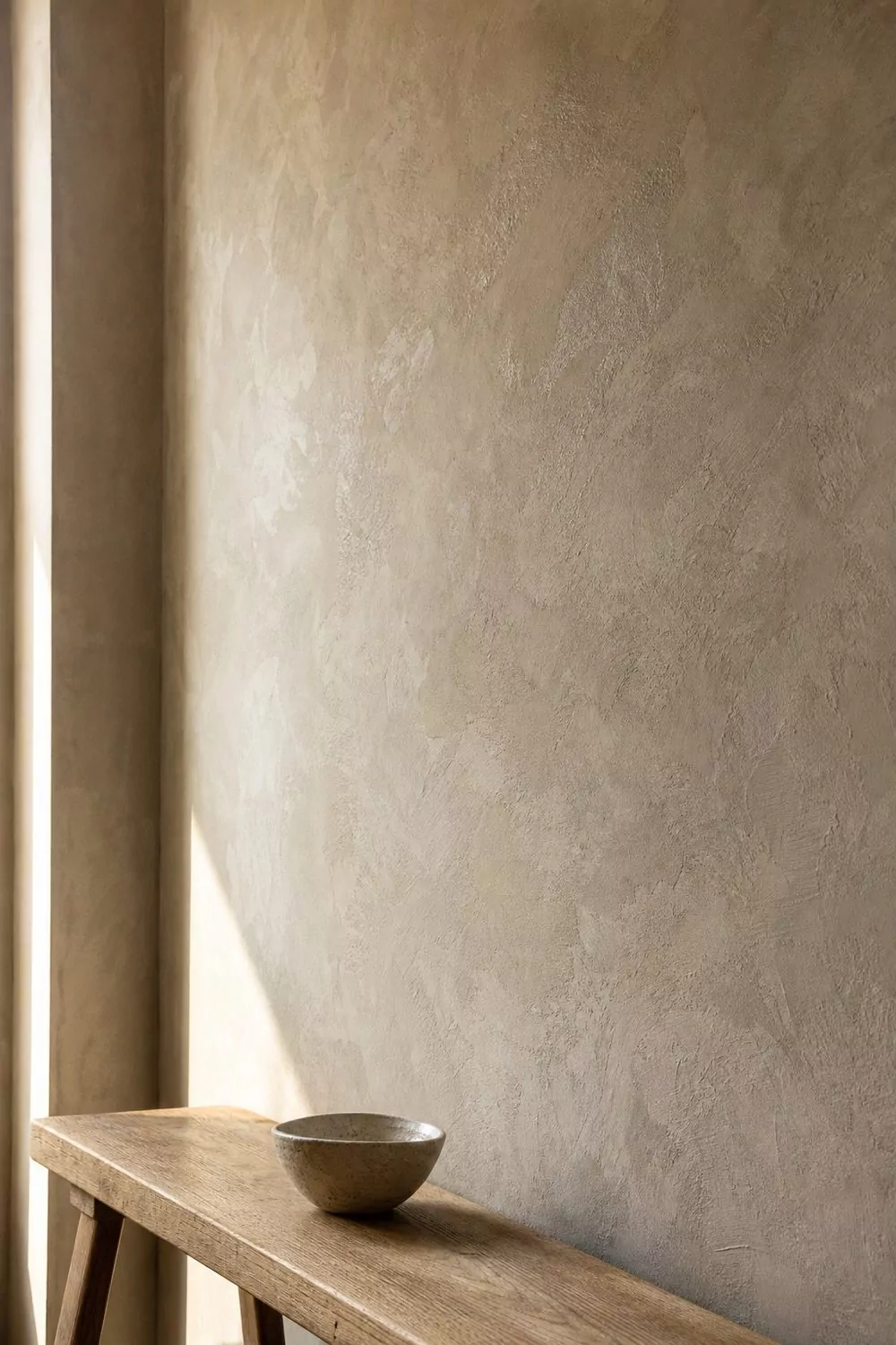 Close view of a beige limewash plaster wall with cloudy tonal variation and trowel texture above a simple oak bench