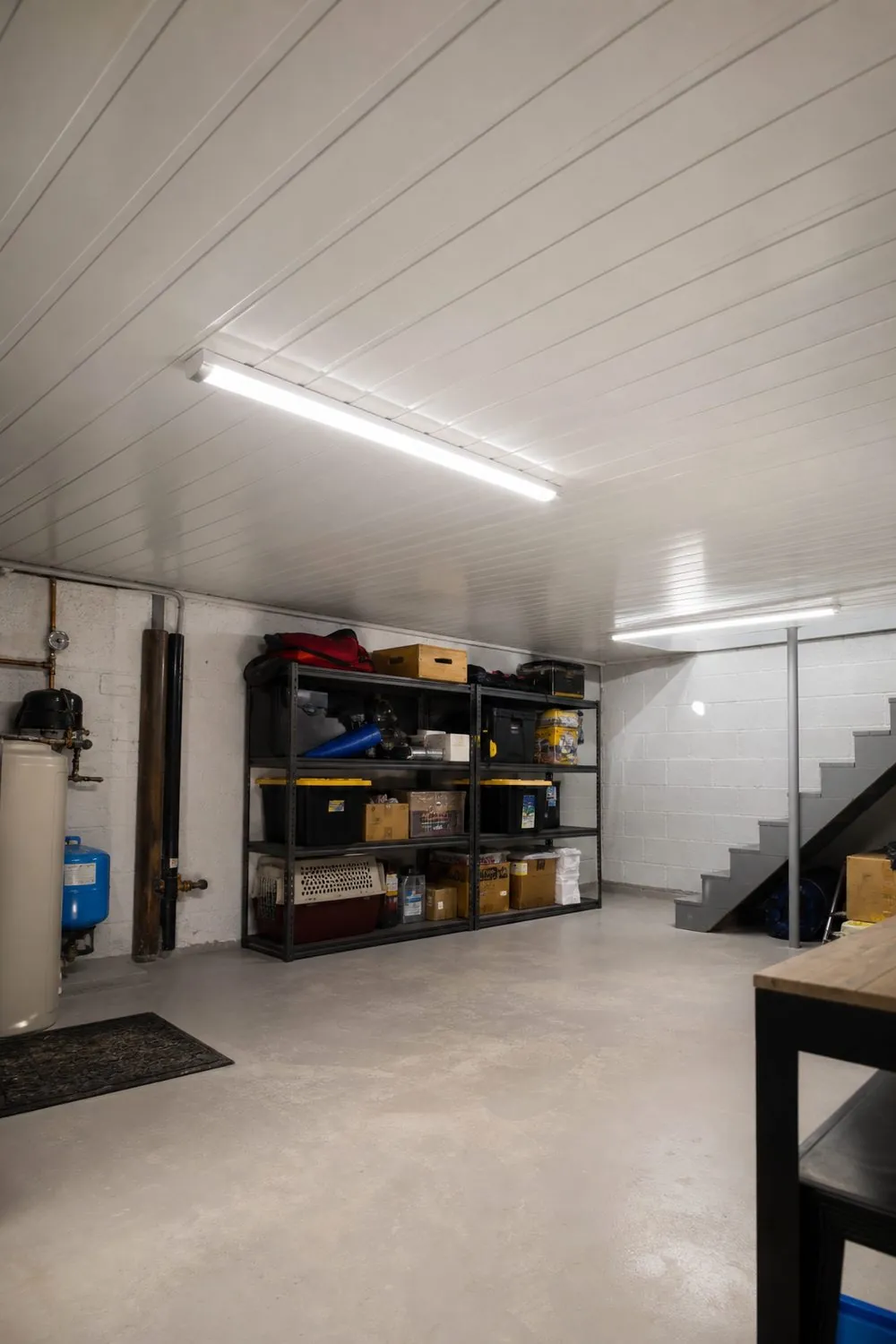 Basement utility room with a bright white interlocking PVC panel ceiling and linear LED shop lights above metal storage shelving.