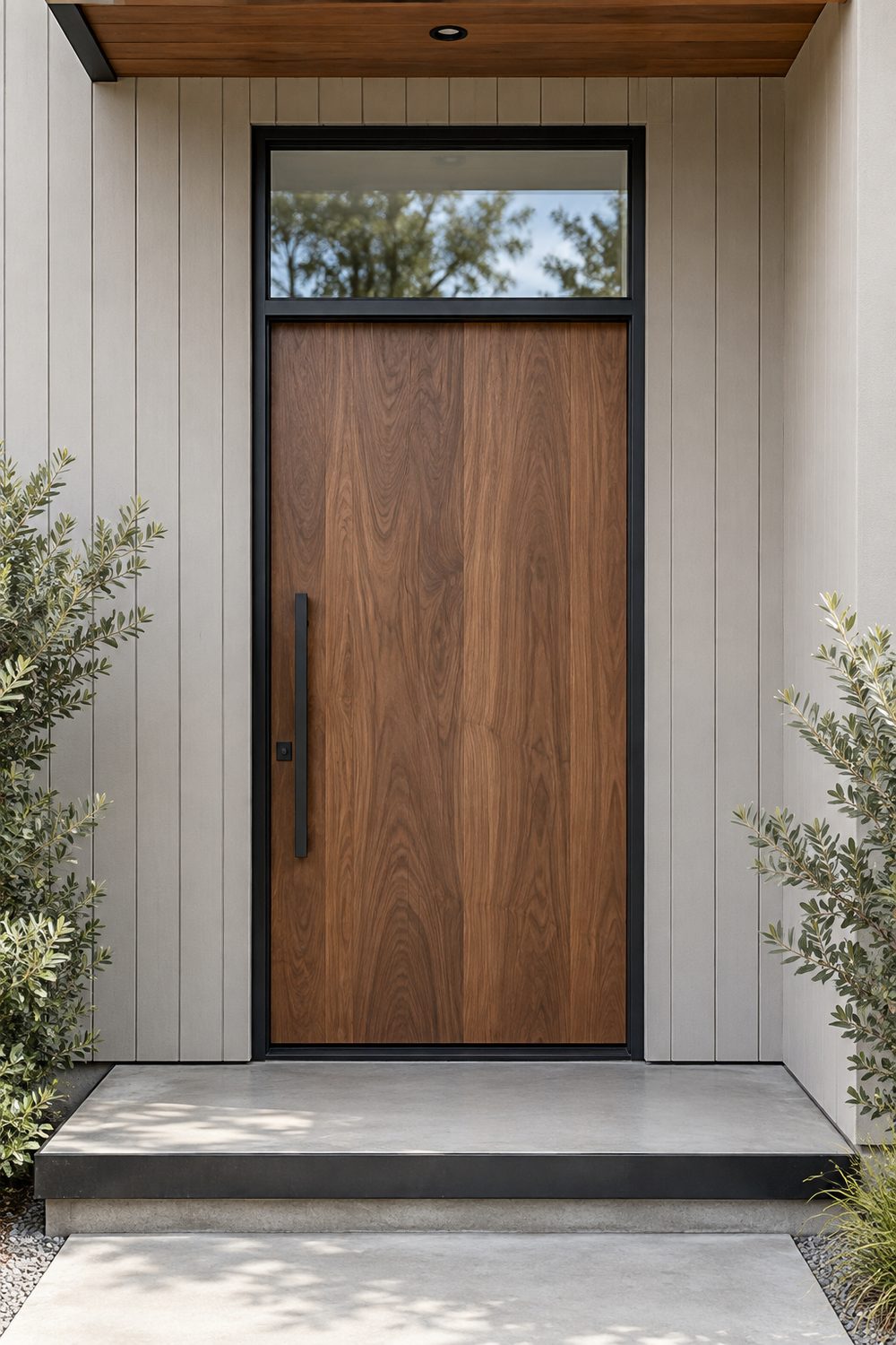 Walnut front door with a full width clear glass transom above in a slim black frame, set in pale grey vertical siding