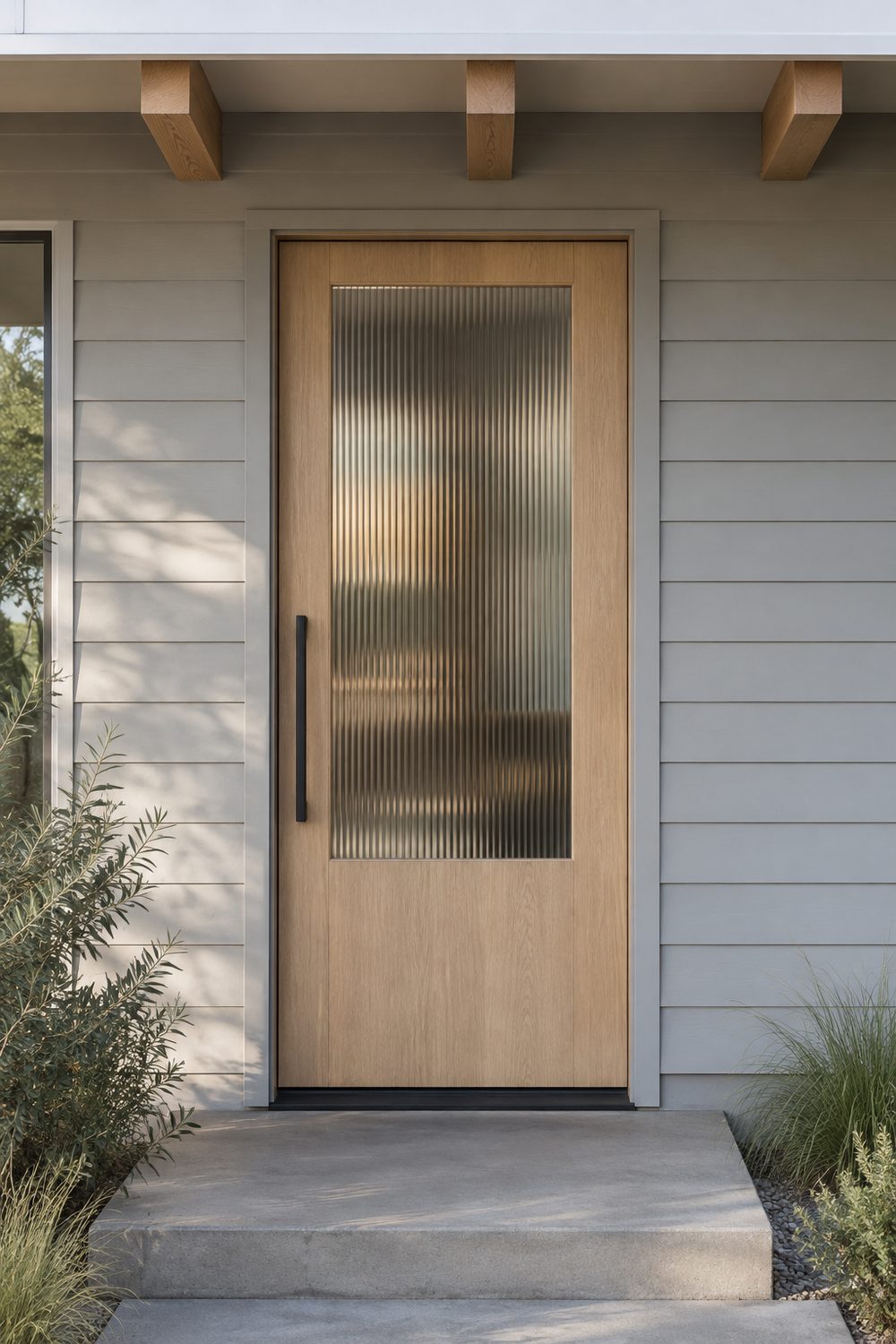 White oak front door with a tall vertical reeded glass panel and slim black pull set in pale grey board siding
